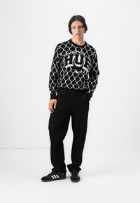 Sort og hvid langærmet sweater med geometrisk mønster og "HUF" logo, parret med løstsiddende sorte bukser og sorte sneakers.