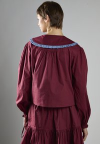 Blusa marrone con maniche a palloncino, colletto blu arricciato e dettagli arricciati sul retro, abbinata a una gonna marrone coordinata.