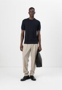 Mornarsko modra pletena majica s kratkimi rokavi, bež nabran hlače, črne čevlje in temno torbo. Model stoji proti enobarvni beli ozadju.