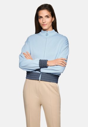 Frau mit langem, dunklem Haar trägt einen hellblauen Pullover mit Reißverschluss, dunklen gerippten Bündchen und Bund, beigen Hosen, steht mit verschränkten Armen.