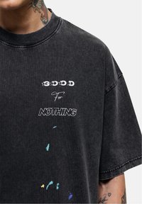 Musta oversize t-paita, jossa on pyöreä kaula-aukko, näyttävä valkoinen graafinen printti ja värikkäitä roiskeita, valmistettu pehmeästä ja teksturoidusta kankaasta.