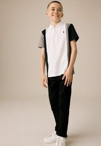 Polo à manches courtes en blanc, gris et noir avec un col zippé et un détail de logo, associé à un pantalon noir et des baskets blanches.