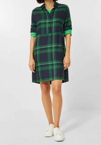 Donna che indossa un vestito a quadri verde e blu navy, lungo fino al ginocchio, con maniche arrotolate e sneakers bianche, in piedi su uno sfondo neutro.