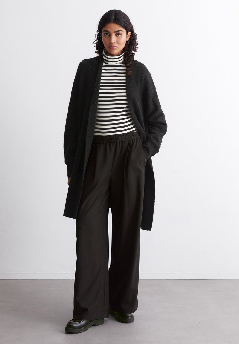 Langer schwarzer Cardigan aus weichem Stoff, über einem schwarz-weißen gestreiften Rollkragenpulli getragen. Lockere schwarze Hose und klobige schwarze Schuhe.