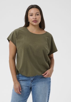 Woman with long dark hair wearing olive green short-sleeve top and light blue jeans, standing against a plain white background.