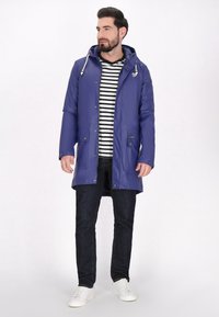 Fioletowa wodoodporna parka z kapturem, z dwoma przednimi kieszeniami i zatrzaskami, noszona na czarno-białą w paski koszulę oraz ciemne dżinsy. Białe sneakersy.