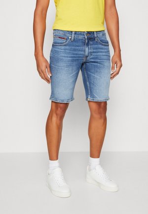 Homme portant un short en jean bleu moyen, des chaussettes blanches, des baskets blanches et un t-shirt jaune, debout devant un fond clair uni.