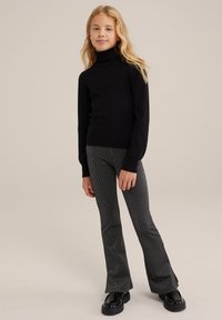 Juodas raštuotas turtlenka su ilgomis rankovėmis, derinamas su pilkų flared tipo kelnėmis, turinčiomis vertikalias juosteles, ir dėvimas su stambiais juodais batais.