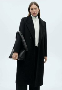 Zwarte wollen jas met een dubbelknopssluiting. Draag het over een witte coltrui en gecombineerd met een zwarte leren handtas. Eenvoudige, moderne silhouet.