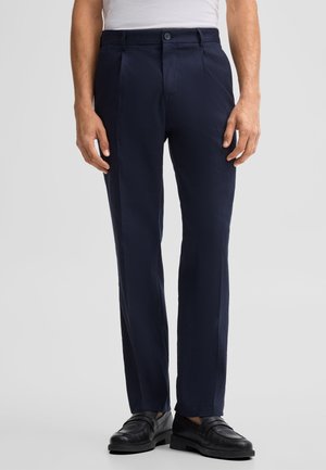 Mann trägt marineblaue, maßgeschneiderte Hose mit Fronttaschen und schwarze Lederloafers auf schlichtem hellem Hintergrund.