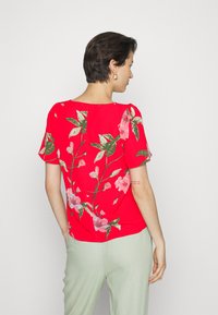 Top vermelho de manga curta com padrões de flores rosa e verdes, decote redondo e um corte descontraído. Combinado com calças verde claras.