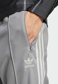 Grå adidas träningsbyxor med dragkedjeficka, med tre vita ränder och en vit logotyp på låret. Slät textur.