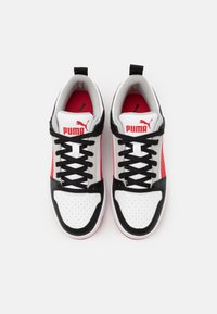 Weiße und schwarze Sneaker mit roten Akzenten, die eine gelochte Zehenpartie, schwarze Schnürsenkel und das Puma-Logo auf Zunge und Seitenpanel aufweisen. Verwendete Materialien mit strukturierter Oberfläche.