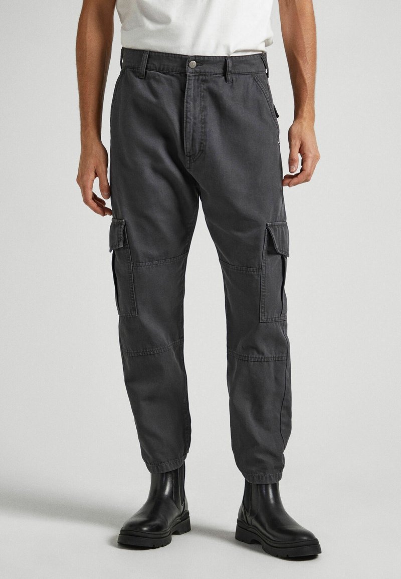 Homme portant un pantalon cargo gris foncé avec des poches latérales et des bottines noires, debout devant un fond clair uni.