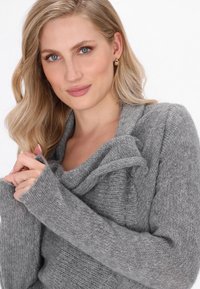 Donna bionda con occhi blu che indossa orecchini a cerchio d'oro e un morbido maglione grigio a maglia, tiene il colletto del maglione vicino al collo.