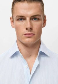 Camisa de botón a rayas azul claro con cuello puntiagudo. Textura de tela suave, corte estándar, con una única tapeta de botones en la parte delantera.