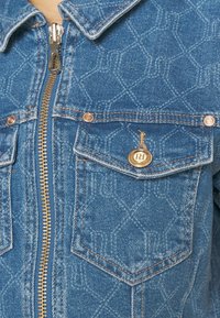 Denimjacka med dragkedja, två framlommer, guldaccenter och ett mönstrat mönster i ljusblått. Stickade detaljer längs sömmarna.