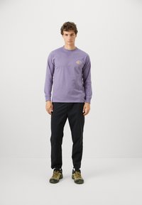 Long-sleeve purple T-shirt with a graphic on the chest, paired with black pants and textured green and black shoes. Model standing.