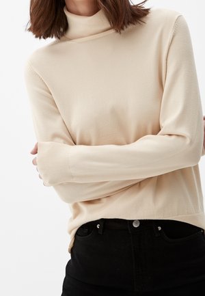 Žena v béžovém roláku s dlouhými rukávy zkřížená přes černé kalhoty s vysokým pasem na bílém pozadí.