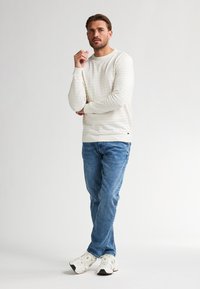 Wit gestreepte trui met ronde hals, gecombineerd met lichtblauwe jeans en witte sneakers. Man staat met gekruiste armen, neutrale achtergrond.