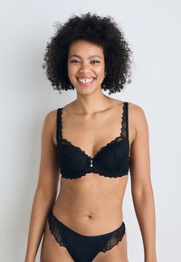 Schwarzer Spitzen-BH mit dekorativem gewellten Rand, Unterstützung durch Bügel und einem kleinen Anhänger in der Mitte. Passende Spitzenunterwäsche ist ebenfalls vorhanden.