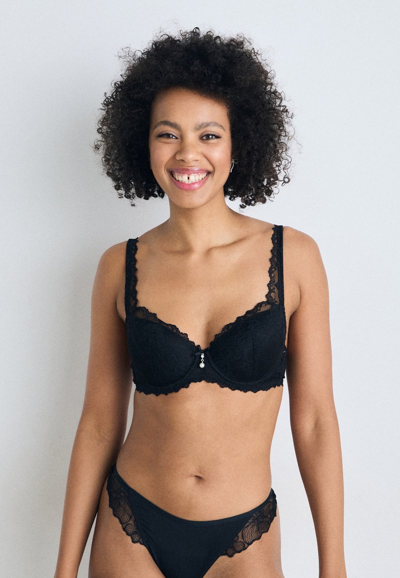 Schwarzer Spitzen-BH mit dekorativem gewellten Rand, Unterstützung durch Bügel und einem kleinen Anhänger in der Mitte. Passende Spitzenunterwäsche ist ebenfalls vorhanden.