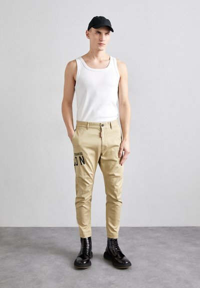 Chinos beige casual con una vestibilità comoda, caratterizzati da una stampa grafica nera e una chiusura con bottone. Abbinati a una canotta bianca e stivali neri.