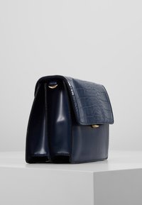 Mørkeblå håndtaske med en blank overflade og krokodillemønsteret klaft, der har en guldfarvet lås og to rum til organisering.