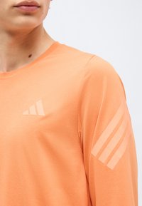 Κοντινό πλάνο ενός ατόμου που φορά πορτοκαλί μπλούζα με μακριά μανίκια, με ένα διακριτικό λογότυπο στο στήθος και ρίγες στο δεξί μανίκι.