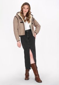 Bruine suede cropped jack met crèmekleurige voering, gecombineerd met een zwarte denimrok met een split aan de voorkant, en bruin knielange laarzen.
