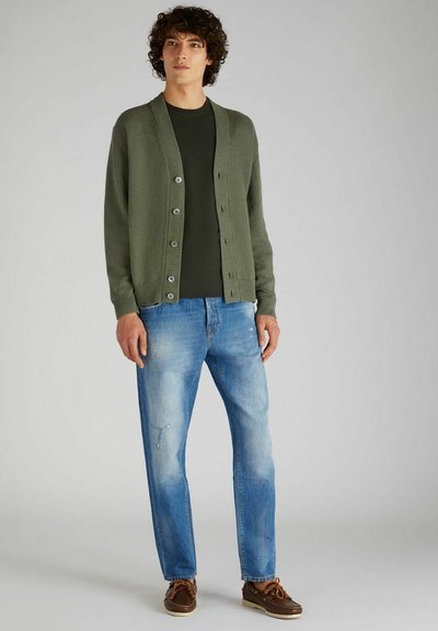 Cardigan verde con bottoni, abbinato a una maglietta verde scuro. Jeans azzurri chiari con leggeri strappi e scarpe da barca marroni.