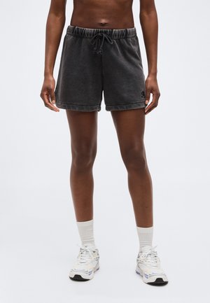 Osoba stojící na jednoduchém světlém pozadí, oblečená v černých krátkých kalhotách se stahovací šňůrkou, bílých ponožkách ke kotníkům a bílých běžeckých botách.