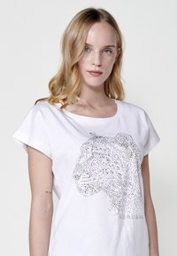 T-shirt en coton blanc à manches courtes, arborant un motif en pointillés de l'Afrique en argent, avec le mot "KOROSHI" imprimé en dessous du graphique.