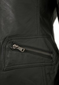 Schwarzes Lederjackenmaterial mit einer Nahaufnahme von einer Reißverschlusstasche. Glatte Textur und sichtbare Nähdetails im Bereich der Tasche.