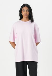Světle růžové oversized tričko s krátkými rukávy a kulatým výstřihem, vyrobené z měkké látky. Nosené s černými kalhotami.