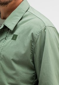 Grüne, leichte Jacke aus glattem Stoff, mit einem Kragen und einem gestickten Logo auf der Brust. Nähte sind sichtbar.