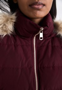 Burgundy vadderad jacka med avtagbar fuskpälskrage, har en silverfärgad dragkedja och horisontella sömdetaljer. Mjukt, texturerat tyg.