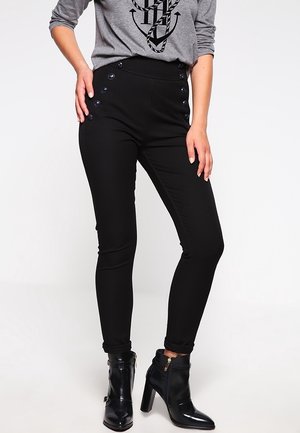 Pantalones negros de corte ajustado con detalles de botones laterales y una textura suave, combinados con una camisa gris de manga larga y botas negras hasta el tobillo.