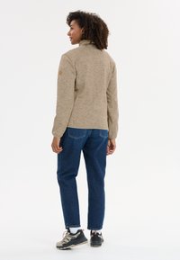 Ljust brunt fleece-jacka med hög krage, prydd med en logotyp-patch på axeln, tillsammans med blå denimbyxor och svarta vandringssandaler.