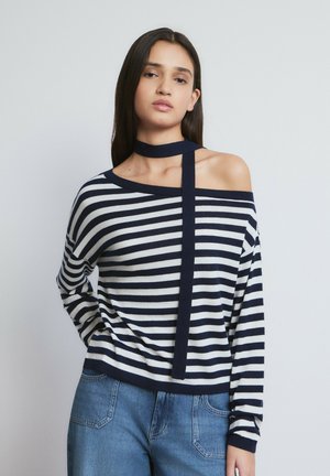 Mujer con cabello largo y oscuro, vestida con suéter de rayas azul marino y blanco, con detalle de lazo en el cuello, y jeans azules, de pie sobre un fondo liso.