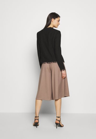 Veste noire texturée avec des franges au bas, associée à des culottes marron amples et des talons aiguilles noirs. Tissu lisse, style ajusté.