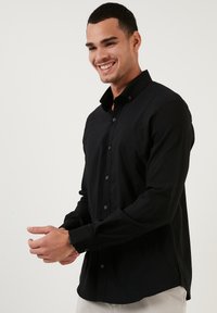 Camisa negra de manga larga con cuello abotonado, de tela texturizada y corte recto. Cuenta con puños abotonados y aberturas laterales para mayor comodidad en el movimiento.
