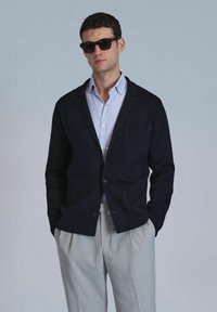Blazer en maille navy avec une légère texture, fermeture à un bouton, deux poches avant, superposé sur une chemise à carreaux bleu clair et un pantalon gris.
