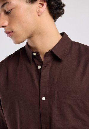 Jonge man met krullend donker haar, die een donkerbruine blouse met borstzak draagt, kijkt naar beneden en opzij tegen een effen achtergrond.