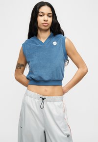 Ženska z dolgimi temnimi lasmi ima na sebi modro brezrokavnico, ki se konča tik nad trebuhom, in ohlapne svetlo sive hlače z vrvico, stoji z rokami na bokih.