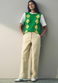 Pull vert à motif argyle sur un t-shirt blanc, associé à un pantalon beige à jambes larges et des baskets vertes et blanches. Fond uni.