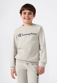 Smilende gutt i lys beige Champion genser og matchende joggebukser, stående mot en enkel hvit bakgrunn.