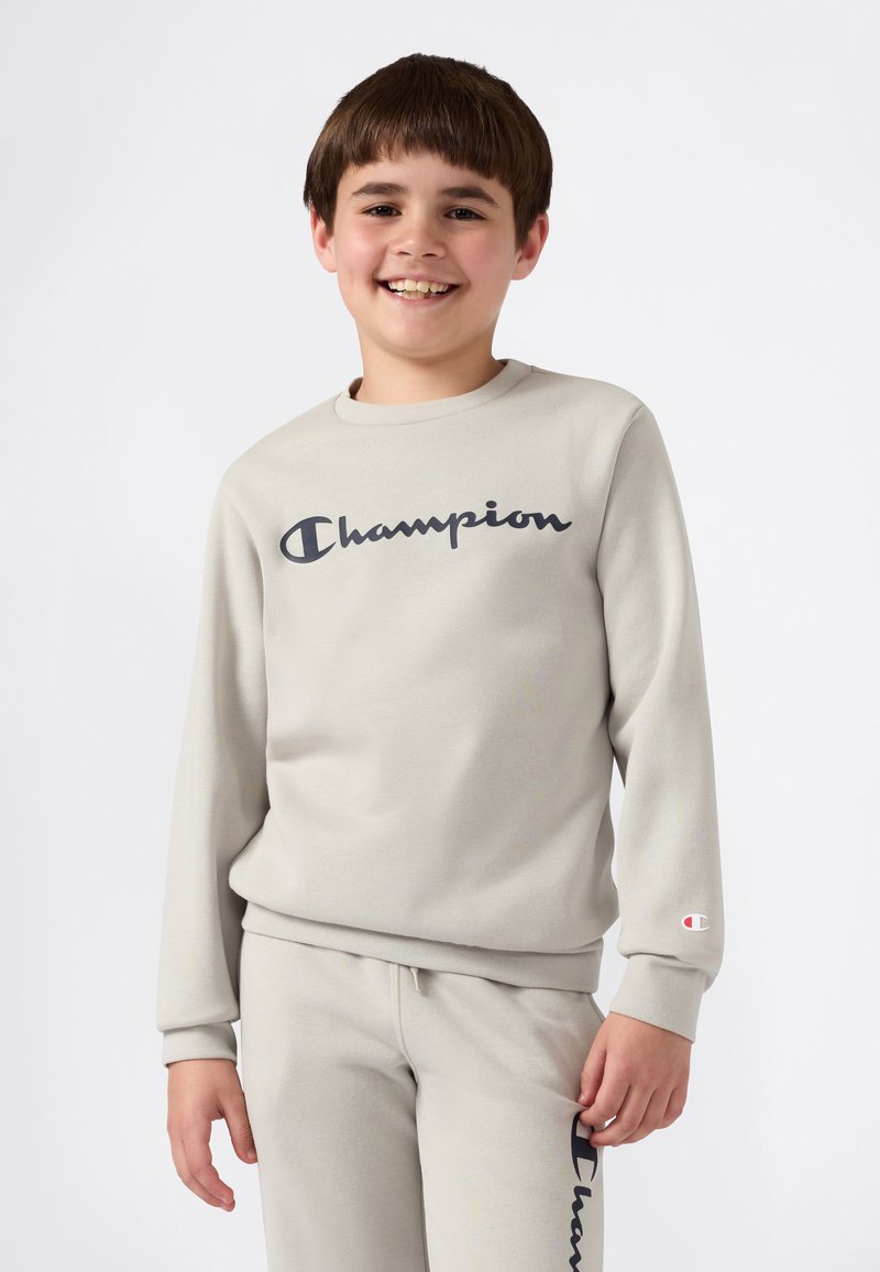 Smilende gutt i lys beige Champion genser og matchende joggebukser, stående mot en enkel hvit bakgrunn.
