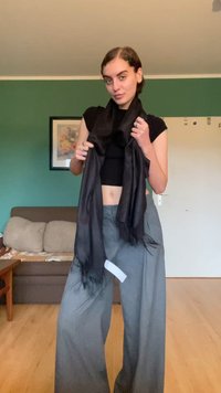 Fular negru cu margini ciufulite, ținută în fața unei persoane îmbrăcate cu un top negru ajustat și pantaloni gri largi. Fundalul prezintă un perete verde.