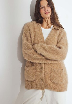 Beżowy, puszysty kardigan o luźnym kroju, z dekoltem w serek, dwoma przednimi kieszeniami i trzema guzikami. Noszony na białym topie, na jasnym tle.